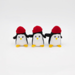 Three small Miniz 3-Pack Penguins with yellow feet, white bellies, and red hats are lined up side by side on a white background.