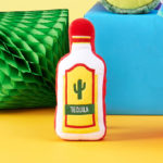Squeakie Pattiez - Tequila in the shape of a tequila bottle with a red cap, yellow label, and green cactus design, set against a yellow and blue background.