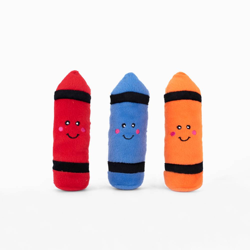 Three plush toys shaped like crayons, colored red, blue, and orange, each with a smiling face and black horizontal stripes near the top and bottom.