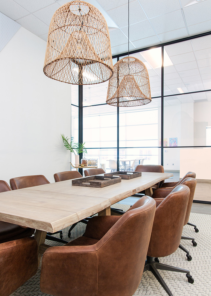 A modern conference room with a wooden table, eight brown leather chairs, large wicker light fixtures, and glass partition walls. A small plant is placed at the end of the table.