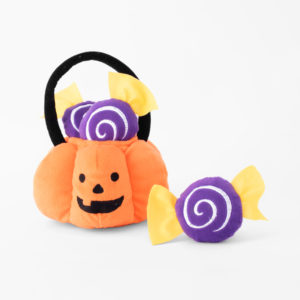 A plush pumpkin basket containing two purple and yellow plush candies, with a third plush candy placed outside the basket, against a plain white background.