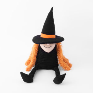 Smiling plush witch doll with orange hair, black cone hat, and black attire, seated against a white background.