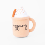 A plush toy in the shape of a beige mug labeled "eggnog," filled with fake whipped cream and sprinkles, with a brown handle and a faux cinnamon stick on the side.