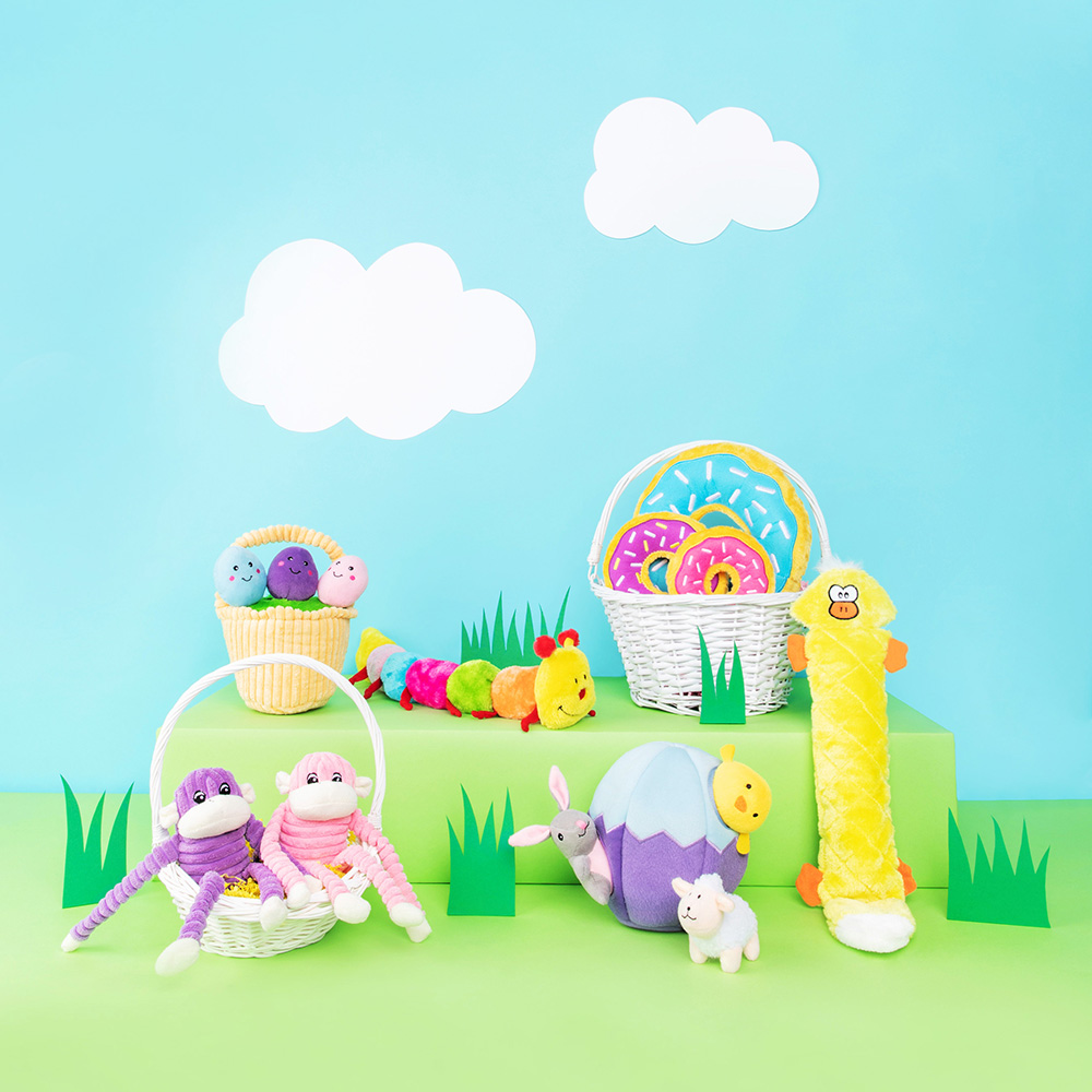 Various colorful soft toys including monkeys, a caterpillar, a chick, eggs, and donuts placed in white baskets and scattered on a green surface with paper clouds in the blue background feature the Easter Miniz 3-Pack Easter Friends.