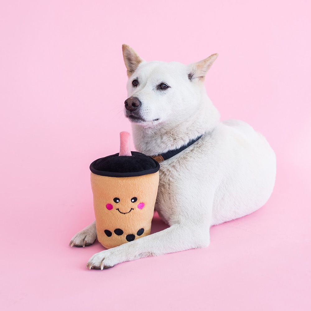 A white dog lies on a pink background, holding a NomNomz® - Boba Milk Tea with a smiling face.