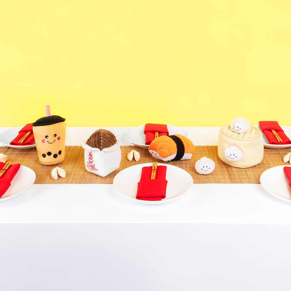 A dining table set with white plates, chopsticks, and red napkins arranged neatly. The table is decorated with plush toys resembling Asian food items, including NomNomz® - Boba Milk Tea, sushi, dim sum, and more.