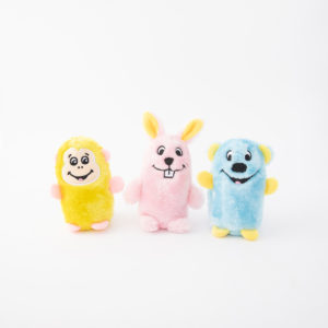 Three plush toys stand side by side: a yellow bear-like toy, a pink bunny with large teeth, and a blue creature with a big smile. The toys are set against a plain white background.