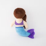 Back view of a stuffed mermaid doll with a brown hairstyle, purple top, and blue tail with purple fin.