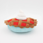 A plush toy resembling a pie with a red lattice crust and a dollop of white topping, set against a plain white background.