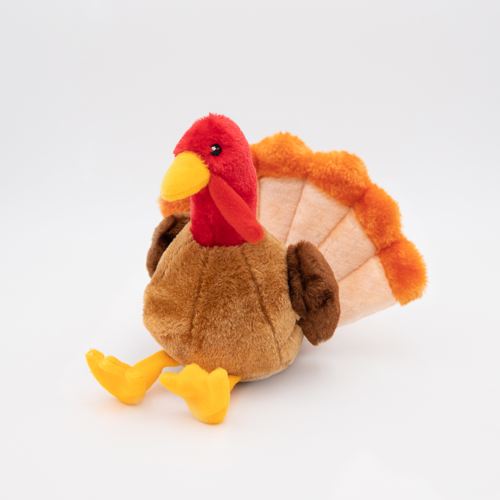 A plush toy turkey with a red head, yellow beak and legs, brown body, and an orange and beige fan-shaped tail, sitting upright on a white background.