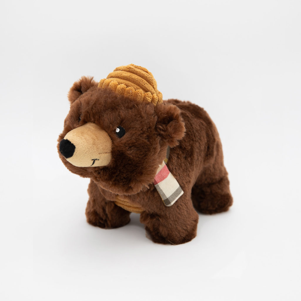 Stuffed brown bear toy wearing a knitted yellow hat and a scarf with a plaid pattern, against a plain white background.
