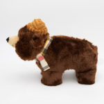 A stuffed brown bear toy with a yellow, red, and beige plaid scarf around its neck, standing on a white background.