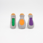 Three soft, plush toy bottles with colored patches (green, orange, and purple) stand side by side against a plain white background.