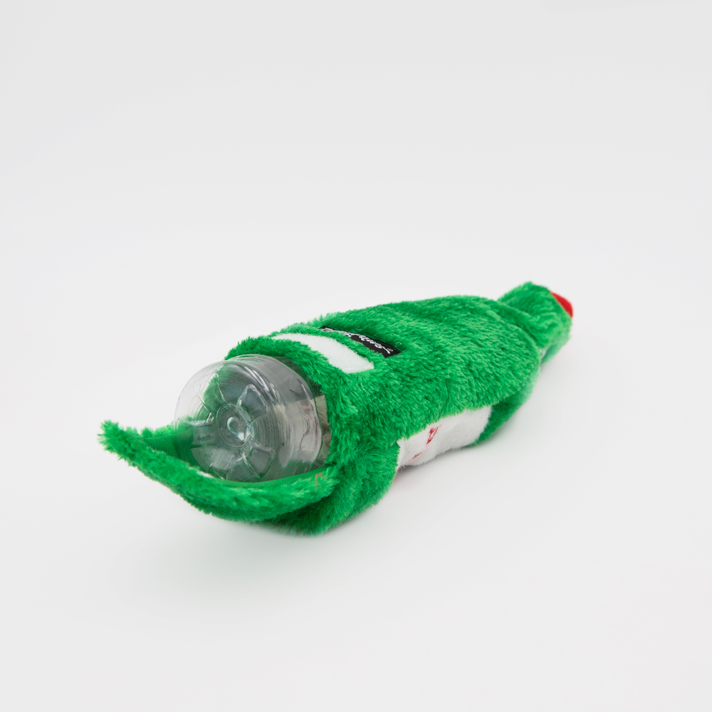 Plush green toy in the shape of a bottle, with a plastic bottle partially visible inside, lying on a white surface.