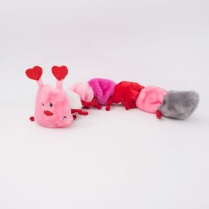 A plush caterpillar toy with segmented, colorful furry sections in shades of pink, red, and gray, smiling face on the front, and red heart-shaped antennae.