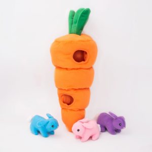 Plush orange carrot toy with three small holes stands upright. A blue, a pink, and a purple stuffed bunny are positioned around it on a white background.