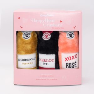 A pink box containing three plush dog toys designed to look like wine bottles. They are labeled "Chardonnay Vintage," "Merlot 2011," and "XOXO Rosé." The box is labeled "ZippyPaws Happy Hour Crusherz.