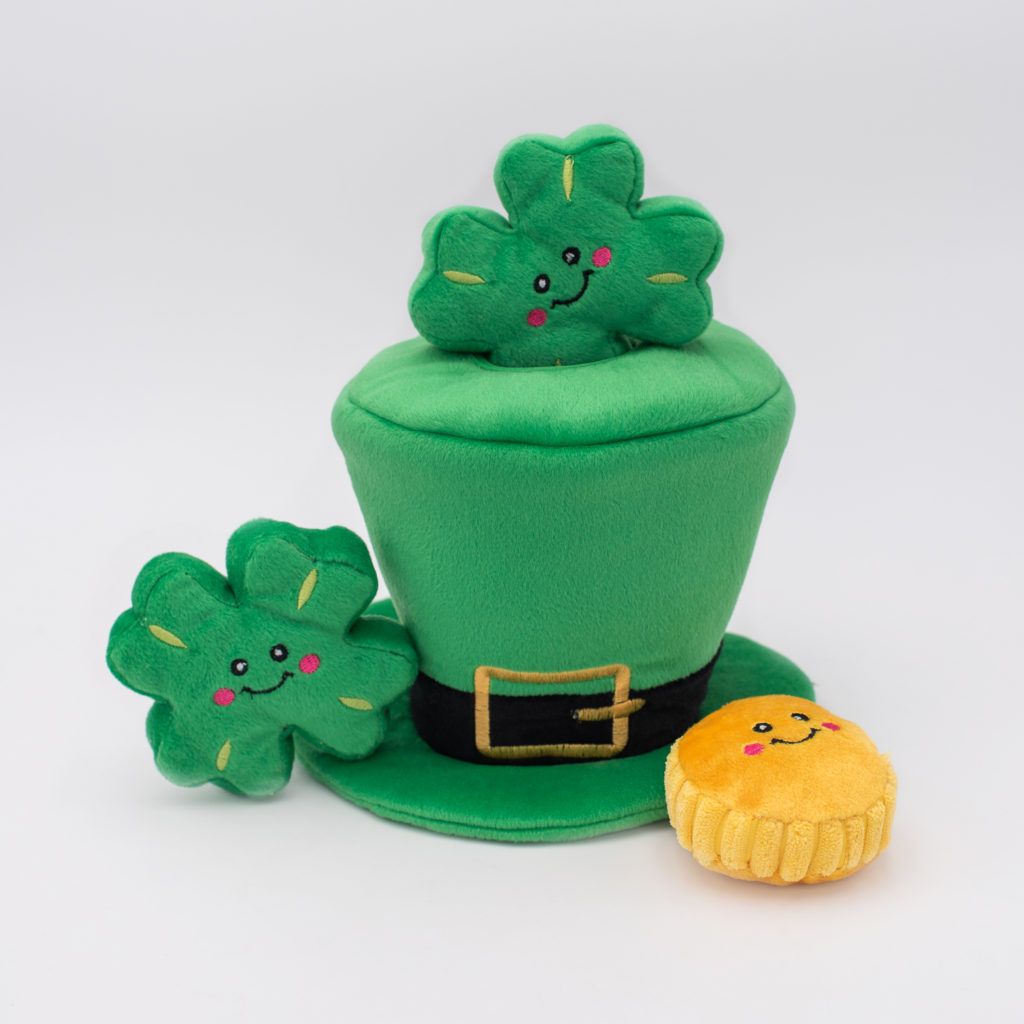 A plush green hat with a black band and gold buckle, topped with two smiling clover toys, and accompanied by a small round yellow plush with a smiling face.