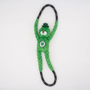 A green, plush bear toy with a cloverleaf on its belly and wearing a small hat is attached to a looped black and white rope, set against a plain white background.