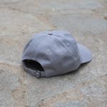 A gray baseball cap lies on a stone surface, shown from the back with the adjustable strap and buckle visible.