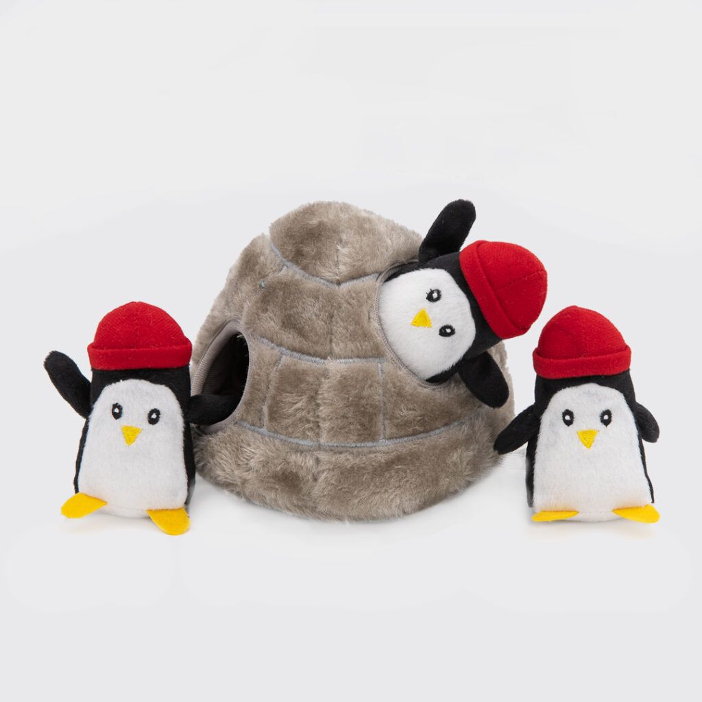 Three plush penguins wearing red hats are positioned around a gray igloo toy. One penguin is partly inside the igloo, while the other two are standing outside.