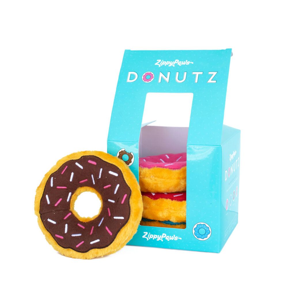 A blue box labeled "Zippy Paws Donutz" containing two plush donut toys and one plush donut toy with chocolate icing and sprinkles displayed outside the box.