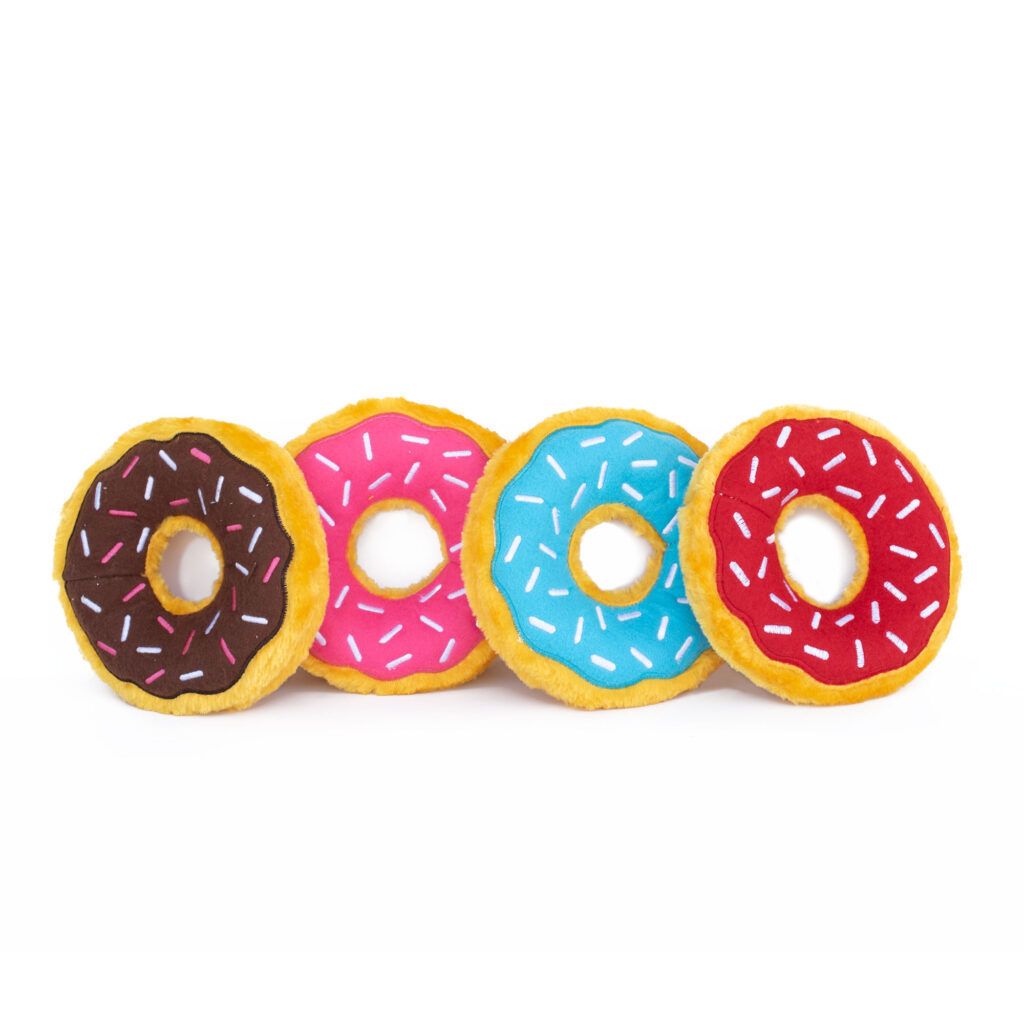 Four plush toy donuts in a row, each with colored icing: chocolate, pink, blue, and red, all decorated with white sprinkles, set against a white background.