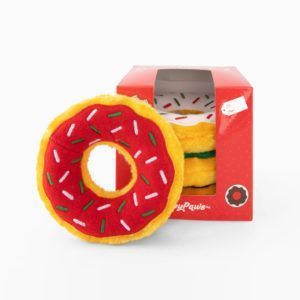 A plush toy resembling a donut with red icing and colorful sprinkles sits next to its red packaging box. The box has a cutout window showing the toy inside.