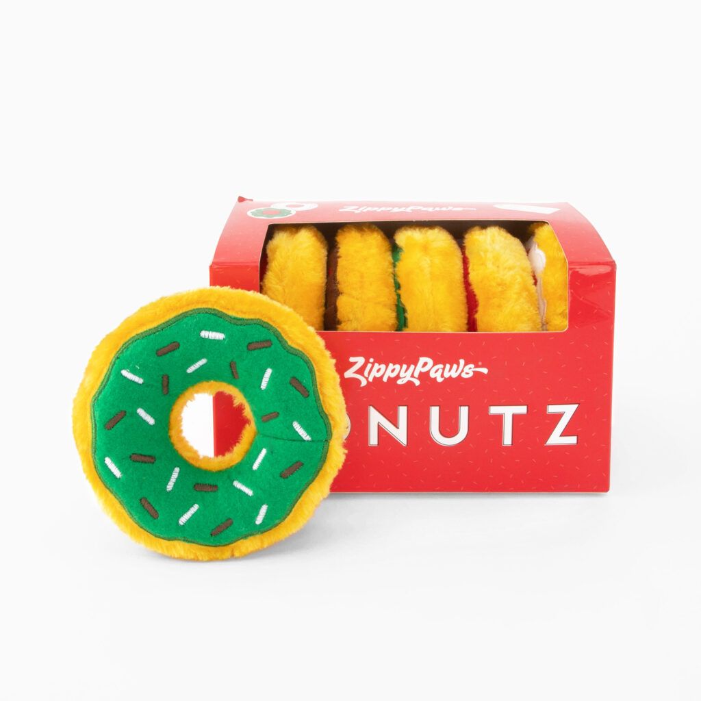 A red box labeled "ZippyPaws Donutz" contains several plush donut-shaped toys with green tops and colorful sprinkles. One plush donut is placed outside the box, displaying its shape and design.