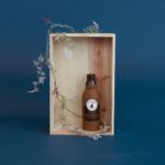 A wooden box contains a brown bottle labeled "Latex Happy Hour Crusherz - Whiskey" with a graphic of a bird. The box is decorated with vines and leaves, set against a blue background.