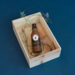 A Latex Happy Hour Crusherz - Whiskey lies inside a wooden crate, placed on a blue surface decorated with small sprigs of greenery.