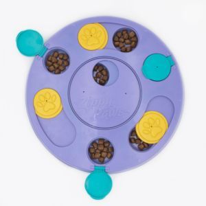 A circular SmartyPaws Puzzler Purple with five compartments, three yellow paw-printed lids, and two teal lids. Each compartment contains brown kibble. The center reads "Zippy Paws.