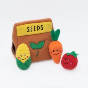 Plush toys including a corn, carrot, and tomato, are arranged in front of a soft, brown bag labeled "Zippy Burrow® - Seed Packet" with a green leaf illustration. Each toy has a smiling face embroidered on it.