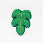 A Squeakie Pattiez - Monstera Leaf shaped like a green leaf with a smiling face and pink cheeks, against a plain white background.