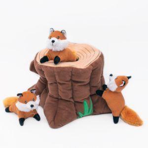 Three Zippy Burrow® - Fox Stump toys are positioned around and inside a plush tree stump toy, with one fox inside the stump, one climbing it, and one sitting beside it.