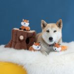 A Shiba Inu dog is laying on a white fluffy surface next to a Zippy Burrow® - Fox Stump against a blue background.
