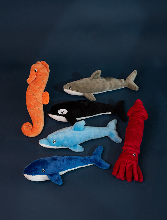 A group of six plush sea animal toys displayed on a dark surface. Includes a seahorse, a dolphin, a whale, a squid, a great white shark, and a killer whale.