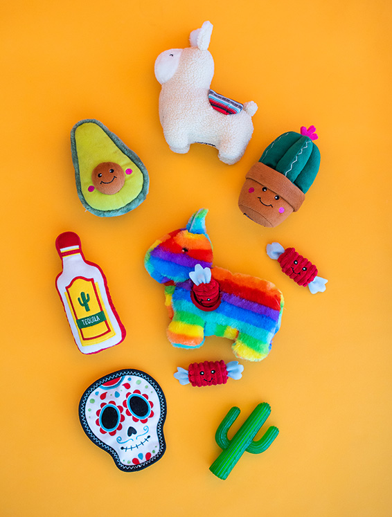 Colorful plush toys including an alpaca, avocado, cactus, pinata, tequila bottle, sugar skull, and wrapped candies arranged on an orange background.