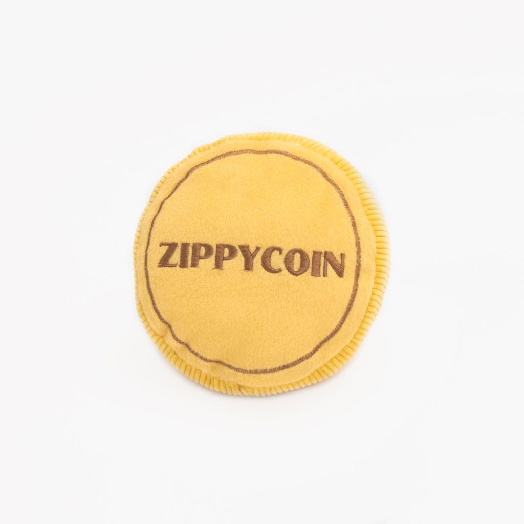 A circular, yellow plush toy with the word "Squeakie Pattiez - Zippy Coin" stitched in brown on its surface.
