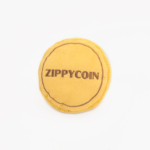 A circular, yellow plush toy with the word "Squeakie Pattiez - Zippy Coin" stitched in brown on its surface.