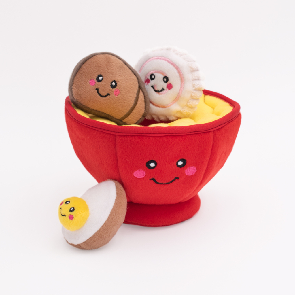 A red Zippy Burrow® - Ramen Bowl filled with smiling plush toys, including a dumpling, a meatball, and an egg, against a white background.