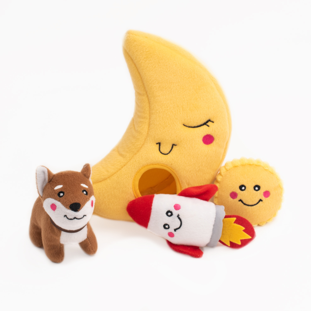 A collection of plush toys including a yellow crescent moon, a brown dog, a red and white rocket, and a yellow sun, all with smiling faces, set against a white background is called the Zippy Burrow® - To the Moon.