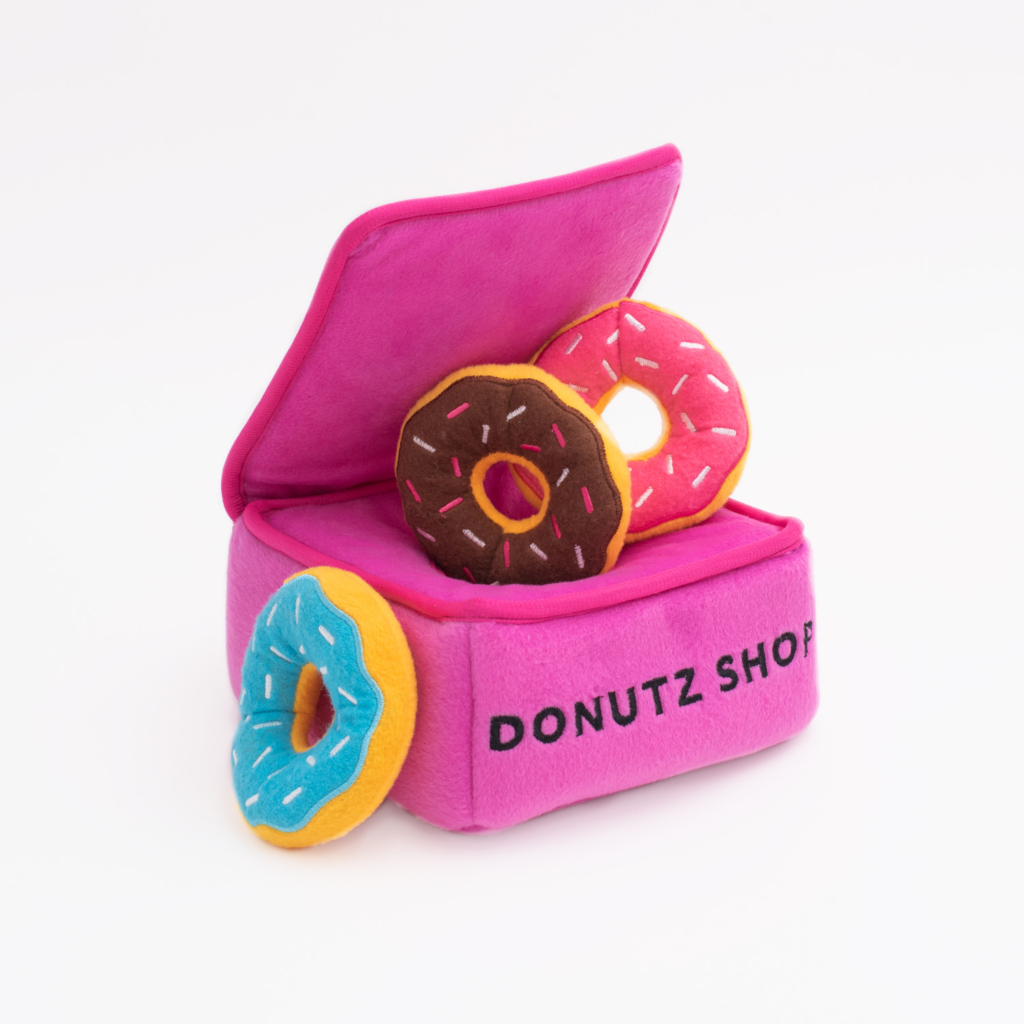 A Zippy Burrow® - Donutz Box shaped like a pink donut shop displays four plush donuts in various colors: pink, brown, blue, and yellow. The words 