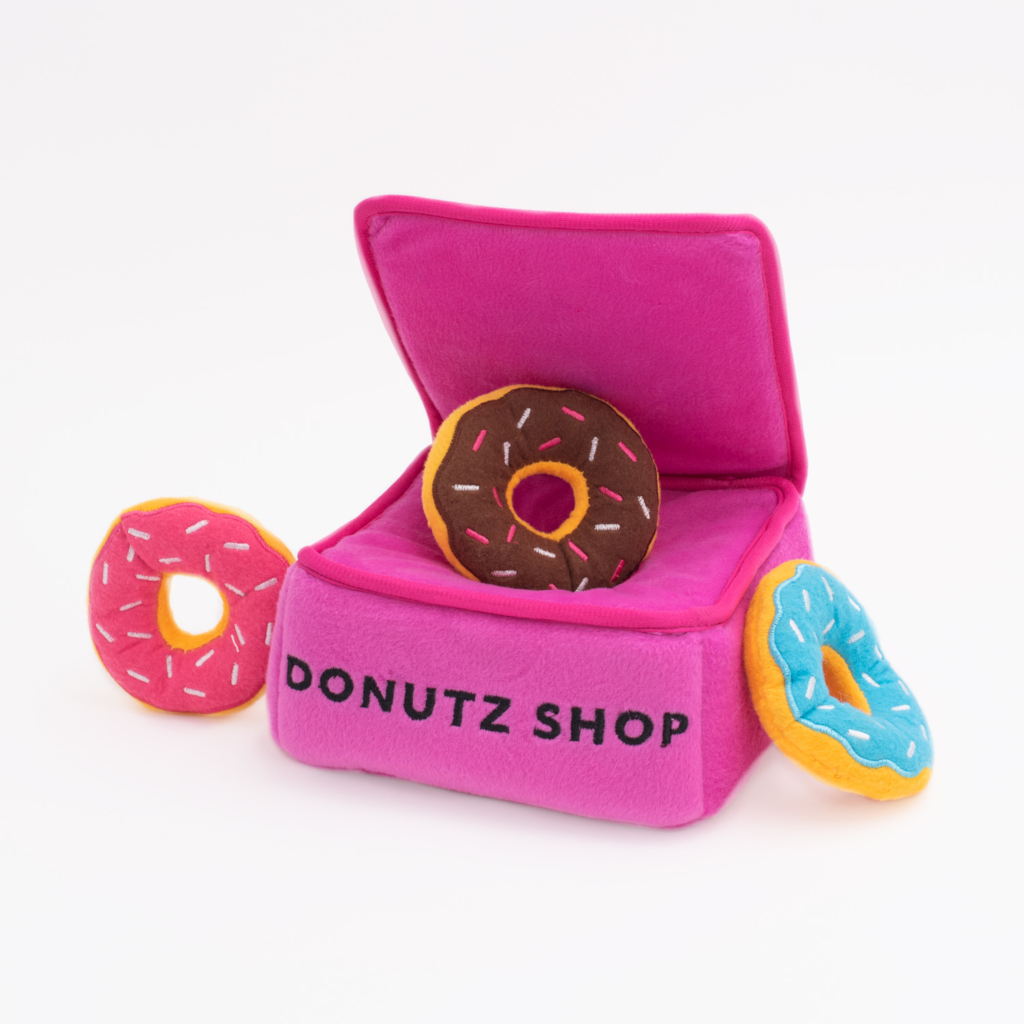 Plush toy doughnuts with sprinkles, in various colors, are placed on and around a pink, fabric box labeled 