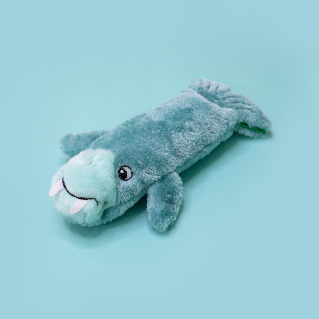 A soft, blue-green Bottle Crusherz - Walrus plush toy with white tusks and black eyes, lying on a light blue background.