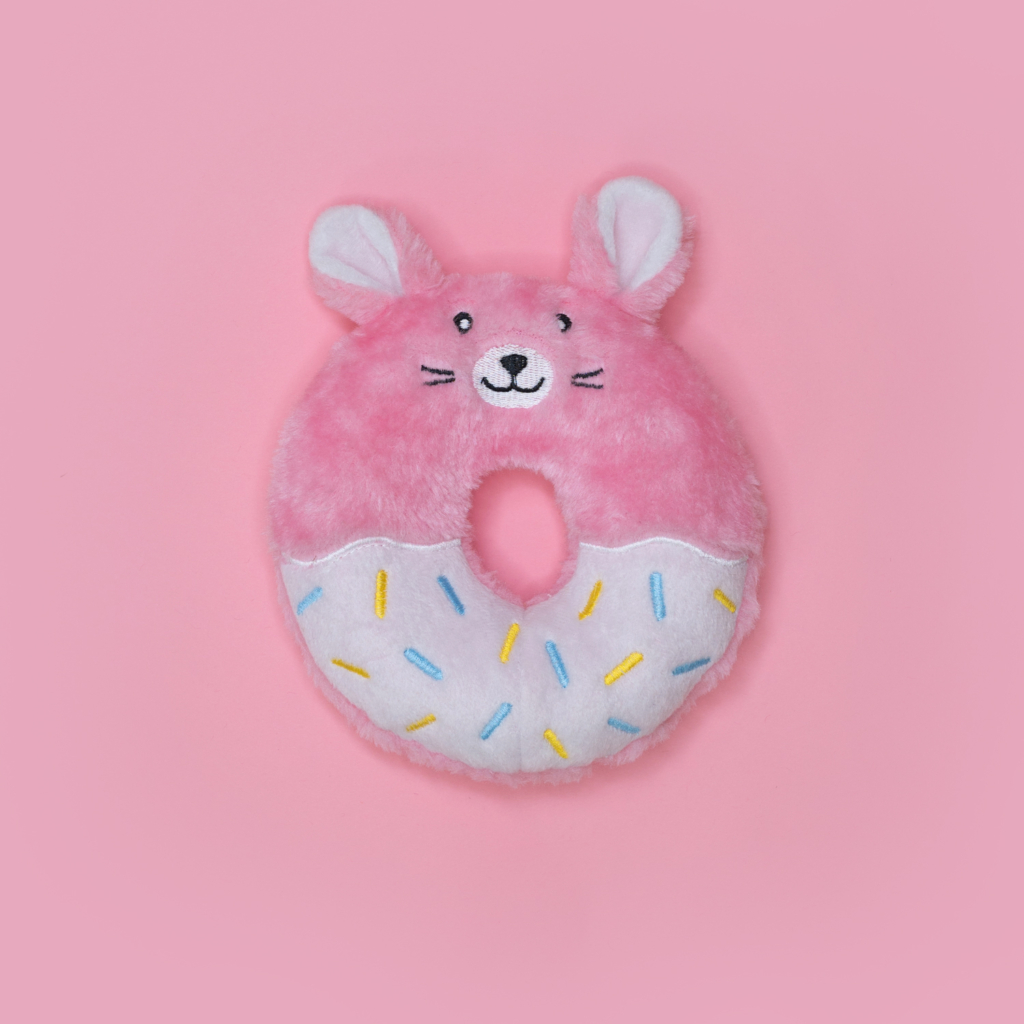 A round, pink stuffed toy shaped like a donut with animal features, including ears and a face, adorned with colorful sprinkles on a pink background called Donutz Buddies - Bunny.