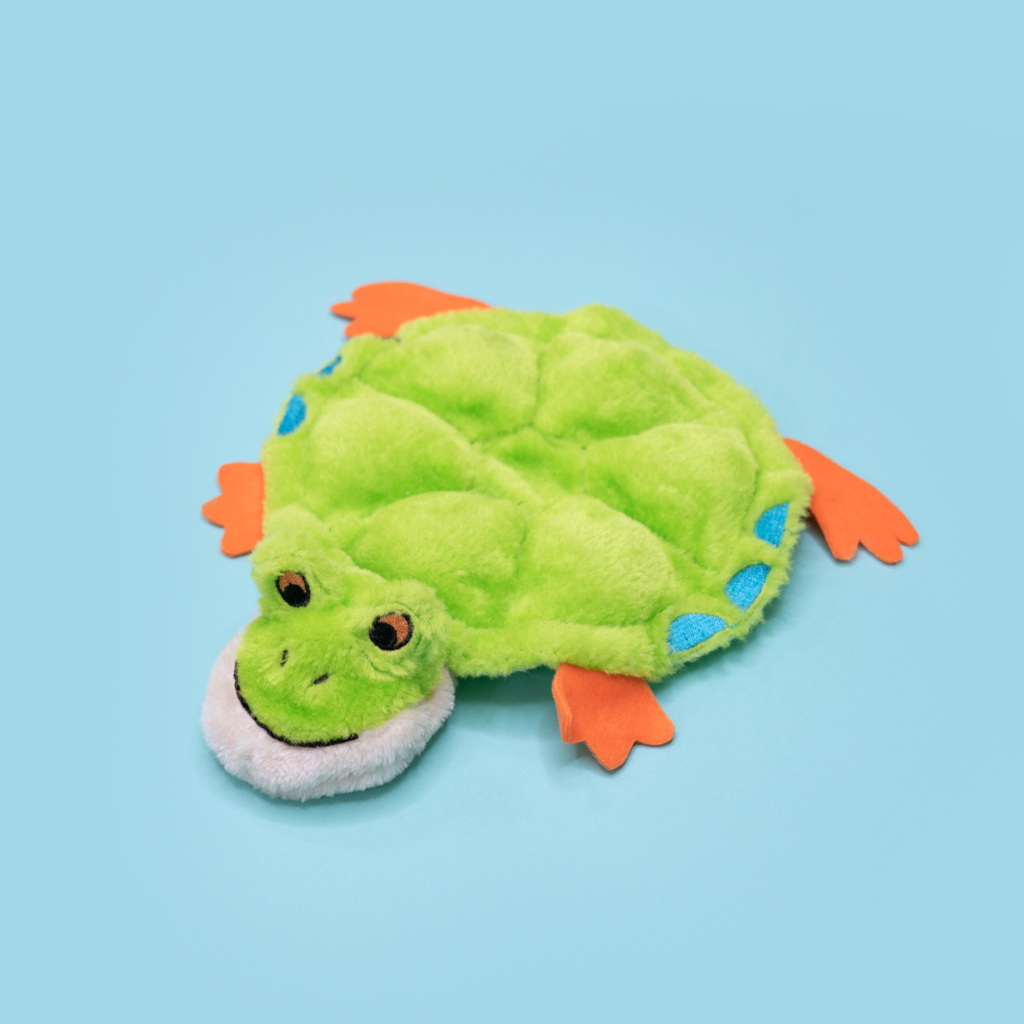 A lime-green plush toy shaped like a turtle with orange flippers and a smiling face, laid on a light blue background has been replaced by the product name 