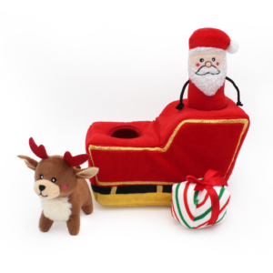 A Holiday Burrow® - Santa's Sleigh next to a small reindeer plush and a white, green, and red striped gift box with a red ribbon.