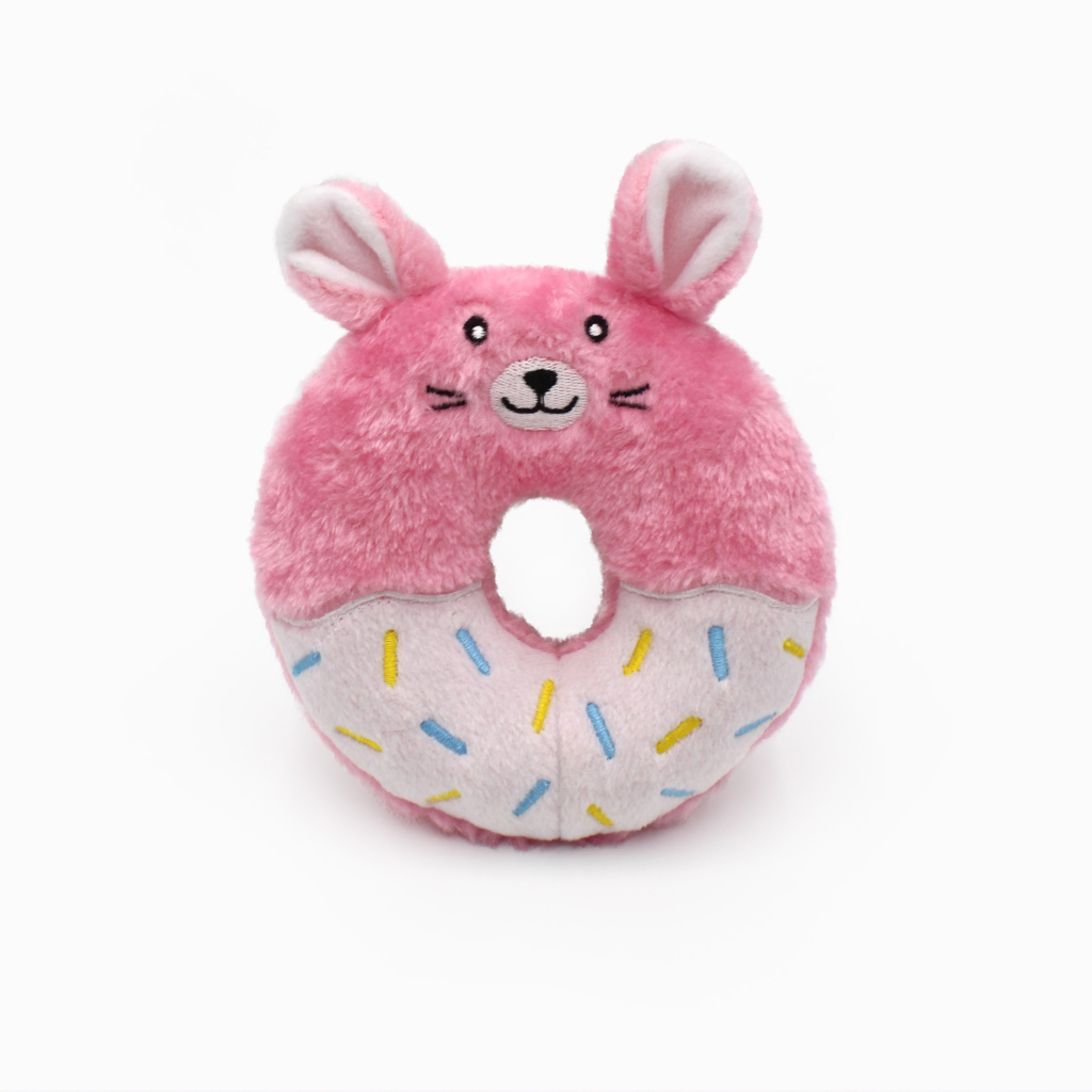 A plush toy shaped like a donut with pink frosting, decorated with multi-colored sprinkles, features a smiling face and bunny ears.