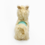 Back view of a plush toy llama with beige fur and a teal saddle.